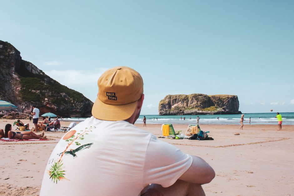 A person wearing a tan cap with 'BRIXTON' embroidered on the back and a white T-shirt featuring a colorful tropical bird and plant illustration sits on a sandy beach, facing towards the ocean. In the background, there are beachgoers sunbathing, walking, and engaging in activities near the shoreline, with small umbrellas, beach chairs, and bags scattered along the sand. The scene is set against a backdrop of rocky cliffs and a clear blue sky, capturing a relaxed seaside atmosphere suitable for outdoor recreation.