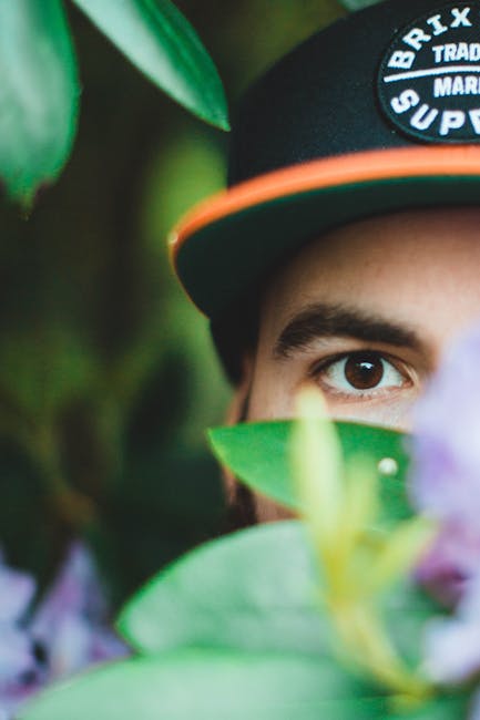 Close-up view of a young man's partially obscured face, showing his brown eye and part of his forehead, as he peeks through green and purple foliage. He is wearing a black baseball cap with orange accents and white text, situated in a natural outdoor setting with soft lighting. The background is blurred with hints of green leaves, emphasizing the foliage surrounding him. The image captures a sense of curiosity and connection with nature, suitable for illustrating themes of environment-friendly or nature-inspired cleaning efforts by Brixton Carpet Cleaning. The focus on the face and vibrant plant life highlights attentiveness to detail and cleanliness in home or venue maintenance contexts, aligning with surface cleaning and sanitisation themes related to the Brixton Academy venue cleaning tips page.