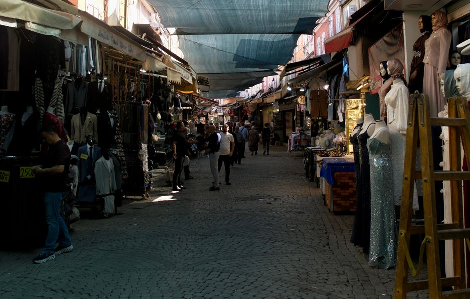 The image depicts a bustling outdoor market scene with a narrow cobblestone pathway lined by various stalls on both sides. On the right, mannequins dressed in elegant clothing and mannequins with headscarves are displayed, along with tables covered in fabrics and accessories. The left side features more market stalls with hanging garments and clothing racks, with a person examining items at one of the stalls. Shade is provided by overhead canopies made of weathered, blue and beige fabric, casting soft shadows over the scene. The sunlight filters through the fabric, illuminating areas of the market while creating contrast with darker regions. The overall cleanliness appears maintained, with no visible dirt or debris on the cobblestone surface. The market is lively, with multiple shoppers walking and browsing the stalls. This scene highlights the vibrant, social atmosphere typical of market environments, where thorough surface cleaning and sanitation are vital to hygiene, aligning with professional cleaning services like those offered by Brixton Carpet Cleaning for commercial spaces. Such deep cleaning helps ensure a hygienic and inviting shopping experience for traders and visitors in SW2, BRIXTON.
