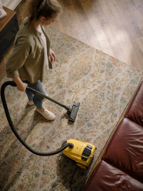 A woman using a yellow vacuum cleaner to deep clean an ornate patterned area rug in a living room, with a brown leather sofa visible to the right and hardwood flooring surrounding the rug. The room is well-lit, highlighting the clean and dust-free surface of the rug, with the woman dressed casually in a beige jacket, gray jeans, and white sneakers, focusing on surface cleaning. The scene illustrates professional domestic cleaning services provided by Brixton Carpet Cleaning, specializing in carpet and area rug sanitisation and maintenance.