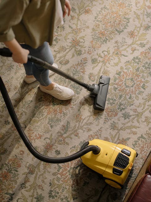 A person vacuuming an ornate, patterned area rug with a yellow carpet cleaner. The individual is dressed in casual clothing, including beige shoes and grey trousers, and is using a handheld attachment connected to a canister vacuum designed for deep cleaning. The room features soft, natural lighting, highlighting the tidy and well-maintained condition of the carpet. This scene illustrates a professional surface cleaning process conducted by Brixton Carpet Cleaning, demonstrating their expertise in residential deep cleaning and sanitisation services in the Coldharbour Lane area of Brixton SW9.