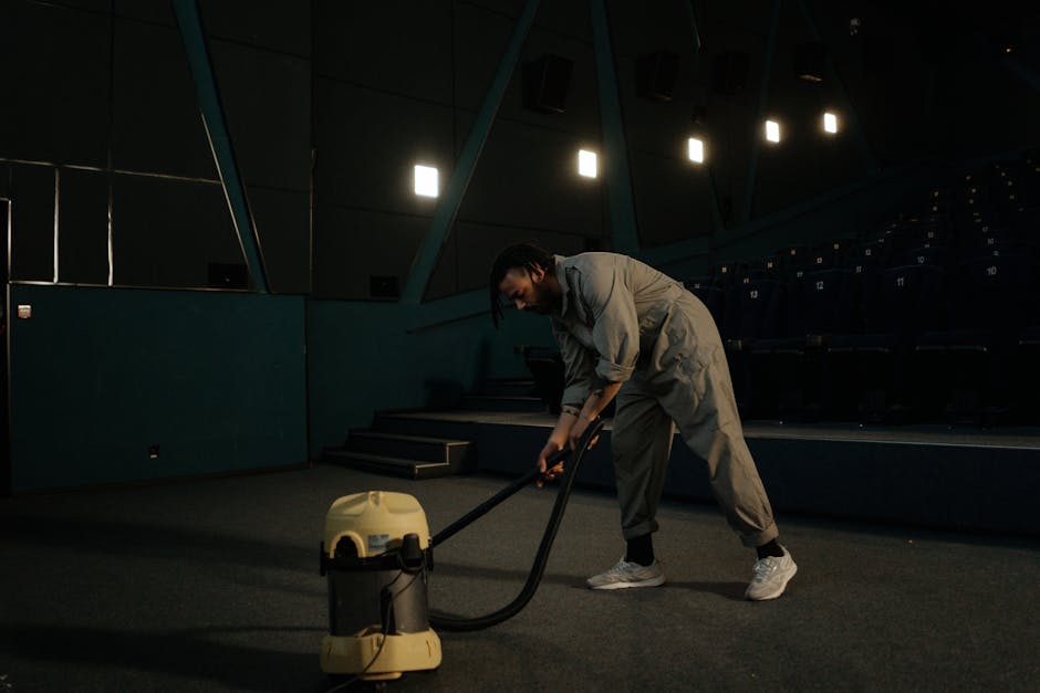 A woman using a yellow vacuum cleaner to deep clean an ornate patterned area rug in a living room, with a brown leather sofa visible to the right and hardwood flooring surrounding the rug. The room is well-lit, highlighting the clean and dust-free surface of the rug, with the woman dressed casually in a beige jacket, gray jeans, and white sneakers, focusing on surface cleaning. The scene illustrates professional domestic cleaning services provided by Brixton Carpet Cleaning, specializing in carpet and area rug sanitisation and maintenance.
