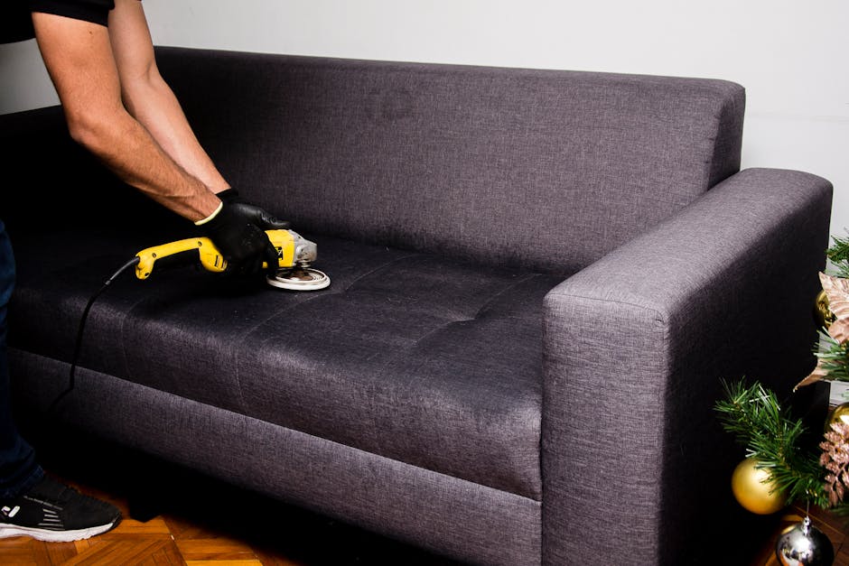 A person wearing black gloves is using a handheld electric fabric steamer to deep clean a dark gray upholstered sofa in a well-lit living room. The sofa features a modern design with wide armrests and a high backrest, and appears to be in a clean, maintained condition. The background includes a white wall and a small Christmas tree decorated with gold and pink ornaments, placed on the right side of the sofa. The lighting highlights the texture of the fabric and the cleanliness of the surface. The scene reflects surface cleaning and sanitisation procedures typical of domestic cleaning services provided by Brixton Carpet Cleaning, focusing on upholstery hygiene.