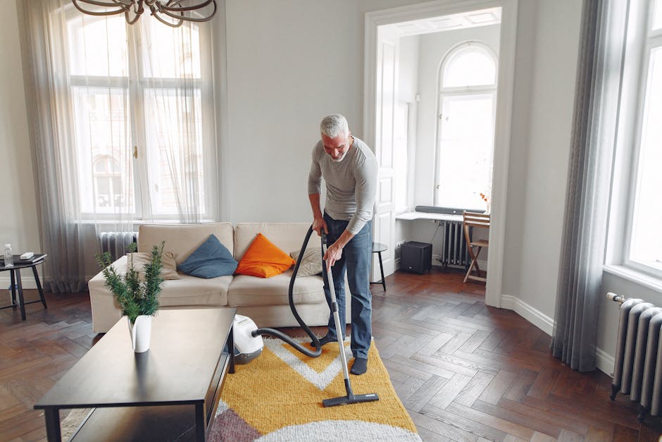 A man with gray hair and glasses is performing surface cleaning in a bright living room, using a vacuum cleaner on a yellow patterned rug. The room features a beige sofa adorned with blue and orange cushions, a dark wooden coffee table with a white vase containing green foliage, and several large windows with sheer white curtains allowing natural light to fill the space. The walls are painted white, and the dark wooden flooring contrasts with the light-colored furniture and curtains. Visible items include a small black speaker, a window seat with a cushion, and a wooden chair near the window. This scene exemplifies deep cleaning and maintenance practices carried out by Brixton Carpet Cleaning, focusing on elevating hygiene standards and preserving the cleanliness of domestic surfaces.