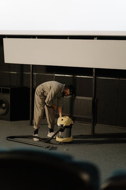 A person dressed in beige overalls operating a yellow industrial vacuum cleaner on a dark-colored carpeted floor in a room with black walls and a large white conference screen or board mounted on the wall. The individual appears to be engaged in a deep cleaning process, focusing on removing dust or debris, with a professional cleaning context. The room features a visible radiator behind the person and audio equipment in the background, indicating a multi-purpose or office setting. Bright lighting highlights the clean and well-maintained environment. Brixton Carpet Cleaning offers such domestic cleaning services, including surface cleaning and sanitisation for commercial spaces.